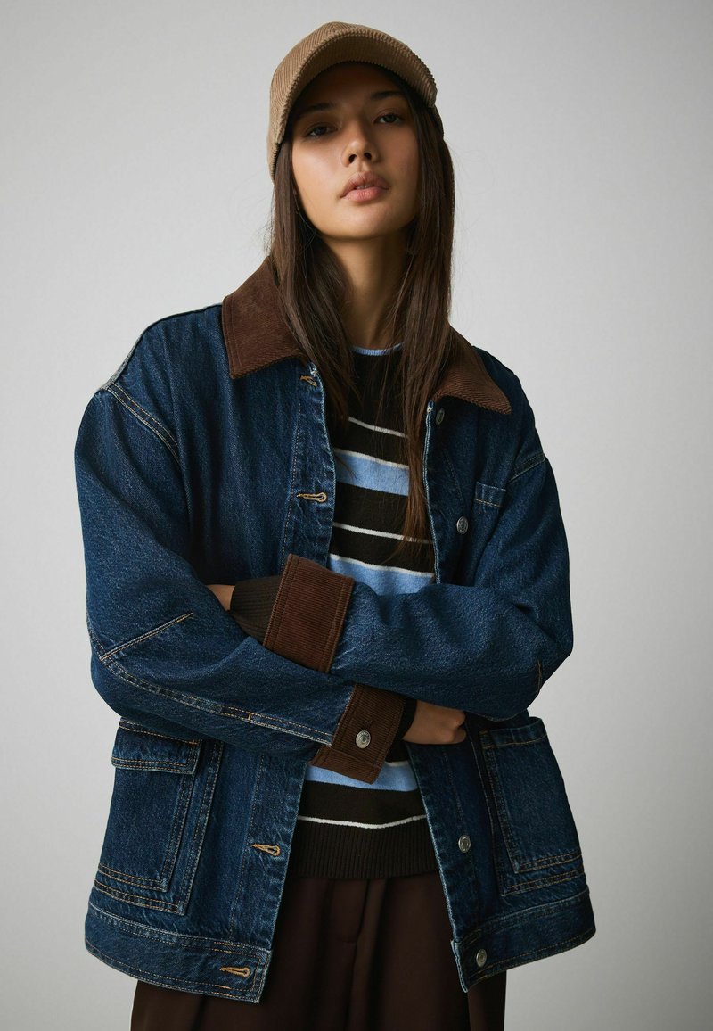 Junge Frau mit langen Haaren, die eine Jeansjacke mit braunem Cordkragen und -manschetten, einen gestreiften Pullover und eine beigen Mütze trägt, die Arme verschränkt.