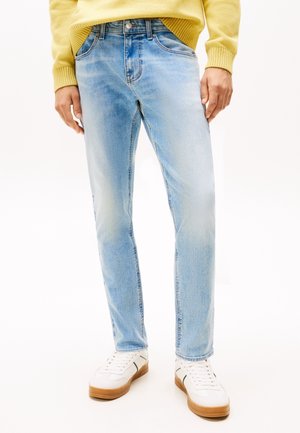 Personne portant un jean bleu clair délavé, des baskets blanches avec des semelles marron et un pull jaune, debout devant un fond blanc uni.