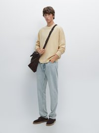 Maglione beige, jeans azzurri chiari e scarpe in camoscio marroni. Borsa a spalla marrone con tracolla. Modella in piedi con un'espressione neutra.