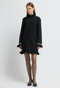 Černé šaty s dlouhým rukávem, volánkovými manžetami a lemem, kulatým výstřihem, jednobarevnou texturou, doplněné o černé ploché boty a průhledné punčochy.