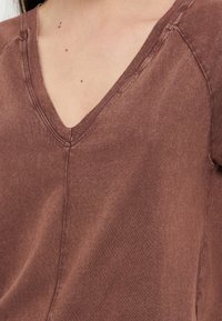 Κοντινό πλάνο σε καφέ πουκάμισο με V-λαιμόκοψη που φοριέται από έναν άνθρωπο, δείχνοντας την υφή του υφάσματος και τις ραφές στο στήθος και τους ώμους.