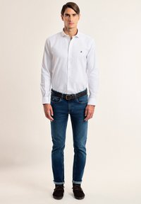Camisa blanca de manga larga con un pequeño emblema, combinada con jeans ajustados azul oscuro y zapatos marrón oscuro. Incluye un cuello abotonado y puños enrollados.