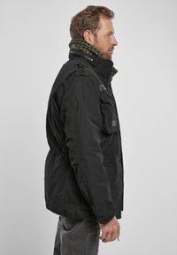 Zwarte stoffen jas met een hoge kraag, meerdere zakken en een verstelbare taille, met daaronder een groen patroonsjaal. Zijaanzicht.