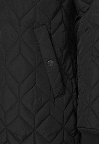 Veste matelassée noire à points losanges, poche à rabat visible avec bouton-pression et poignet côtelé sur la manche.