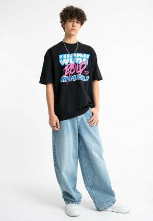 Nastolatek z kręconymi włosami, ubrany w czarną koszulkę z grafiką, luźne jasnoniebieskie dżinsy, białe trampki i srebrny łańcuszek na szyję.