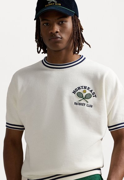 Hvit strikket genser med korte ermer, med marineblå striper på halsen og mansjettene; har et brodert design av kryssede tennisracketer og tekst.