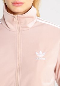 Ljusrosa zip-up jacka med hög krage, med vita randiga detaljer på axlarna och en broderad vit Adidas-logga på bröstet.