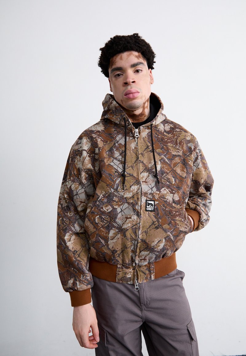Camouflaget zip-up hoodie in bruin en beige tinten, met een bruine geribbelde zoom en manchetten, en een logo-opdruk op de voorkant.