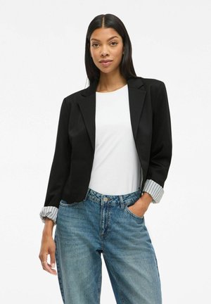 Schwarze Blazer mit tailliertem Design, ausgestattet mit gestreiften Bündchen. Kombiniert mit einem weißen Oberteil und blauen Jeans. Glattes Material und maßgeschneiderte Form.