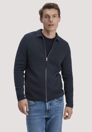 Junger Mann, der einen marineblauen Reißverschluss-Pullover über einem dunklen Hemd und blauen Jeans trägt, stehend vor einfachem hellem Hintergrund.