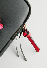 Čierny umeloleatherový puzdro s textúrovaným povrchom, zlatým zipsom a akcentmi v červenej a čiernej farbe. Obsahuje vyšitý ružový dizajn a branding.