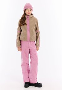 Beige fleece jack met roze accenten, ritssluiting aan de voorkant en capuchon. Lichtroze sneeuwbroek, zwarte laarzen. Model staat tegen een witte achtergrond.