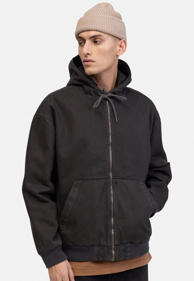 Black hooded jacket made of cotton blend, featuring a front zipper, adjustable drawstring, and two side pockets. Textured, casual fit.