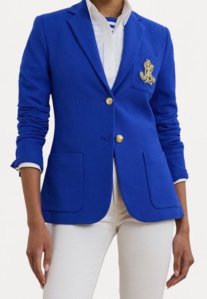 Frau trägt einen blauen Blazer mit goldenen Knöpfen und einem gestickten Wappen, weißes Hemd und weiße Hose, steht mit entspannten Armen.