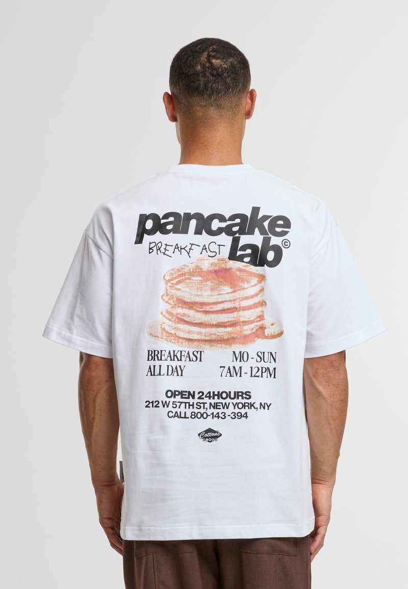 Tricou alb din bumbac cu un grafic reprezentând clătite și text. Designul include un text negru care indică "laboratorul de clătite" și detalii de locație.