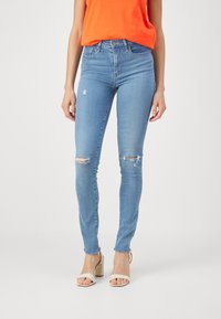 Jeans ajustados de denim azul claro con desgastes y rasgaduras en las rodillas. Cintura alta con un leve deshilachado en el dobladillo. Combinados con tacones beige.