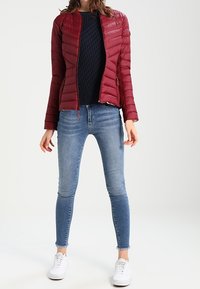 Punainen pehmustettu takki, jossa on muotoiltu design, yhdistettynä tummansiniseen neuleeseen ja vaaleansiniisiin slim fit -farkkuihin. Valkoiset lenkkarit viimeistelevät asun.