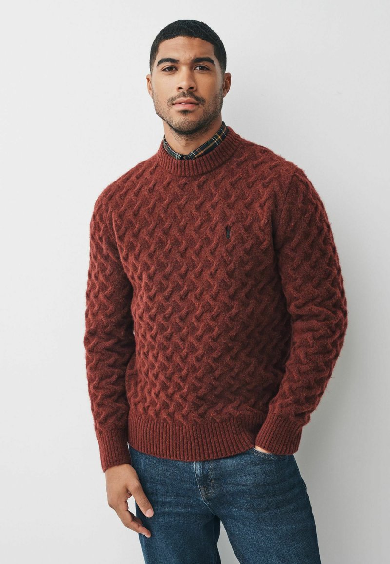 Maglione spesso in rosso scuro, con un motivo a diamante testurizzato, colletto e polsini a costine, abbinato a una camicia a quadri con colletto indossata sotto.