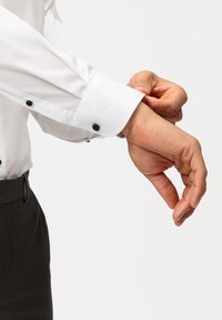 Camicia bianca a maniche lunghe con bottoni neri, dotata di un dettaglio ai polsini. Il tessuto appare liscio con una leggera lucentezza, aderente al polso.
