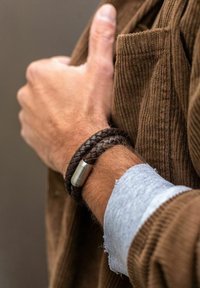 Bracelet en cuir tressé marron avec un fermoir métallique, porté sur un poignet, à côté d'une veste en velours côtelé marron et d'un poignet de chemise bleue.