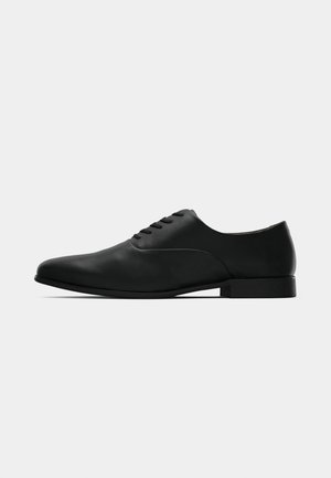 Chaussure oxford pour homme en cuir noir avec un laçage fermé, talon bas et finition lisse et élégante, présentée en profil sur fond blanc.