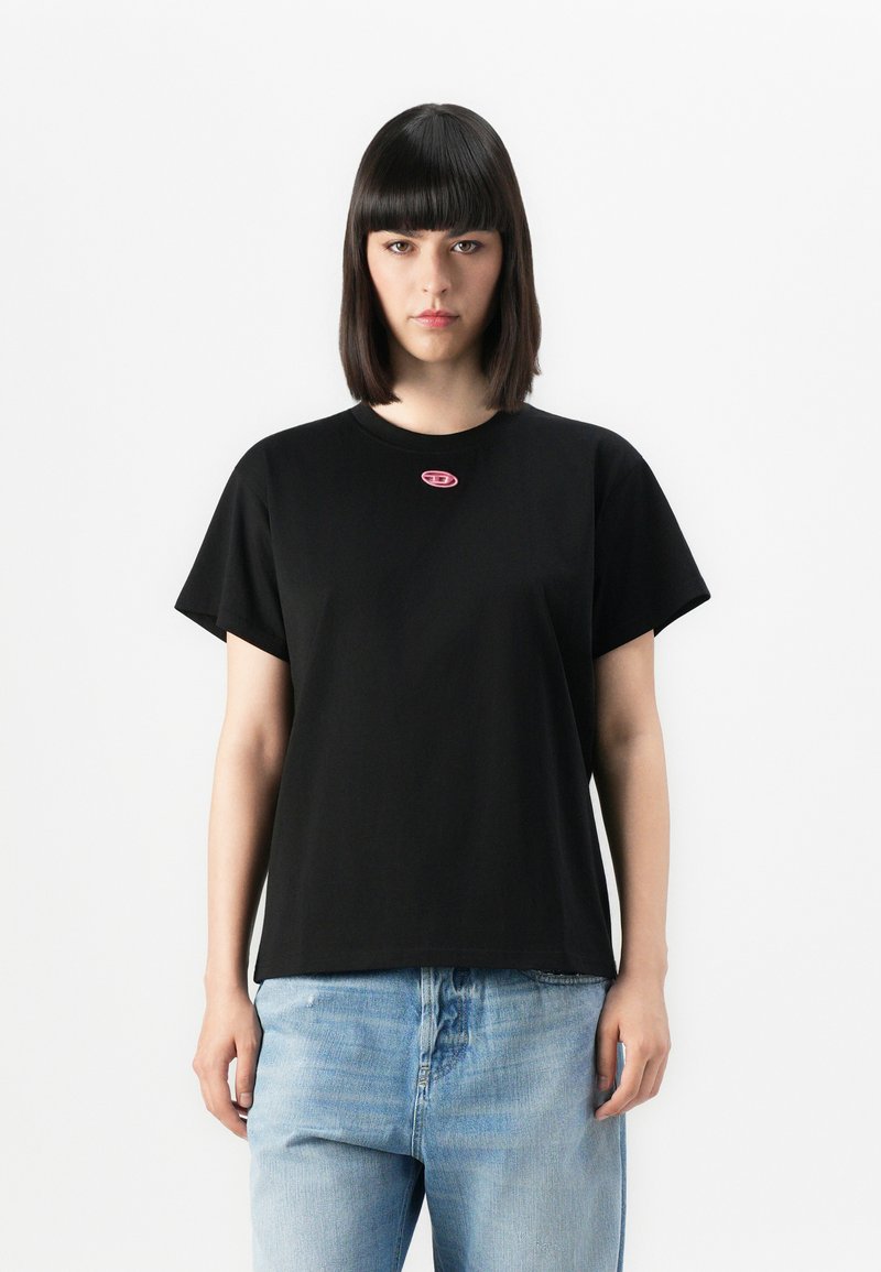 Young person with straight black hair wearing a black t-shirt with a small pink logo and light blue jeans, standing against a plain background.