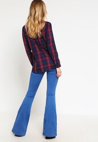 Chemise à carreaux marine avec des accents rouges, assortie à un jean bleu évasé. Tissu lisse, col rond et poignets à boutons visibles.