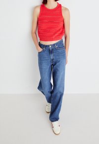 Rode gehaakte crop top met horizontale patronen, gecombineerd met hoge blauwe denim jeans. Witte sneakers met rode accenten maken de look compleet.