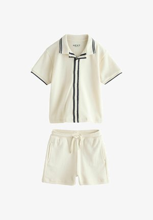 Camicia crema a maniche corte con bottoni e rifinitura nera, abbinata a pantaloncini con coulisse e tasche laterali, disposti su uno sfondo bianco.