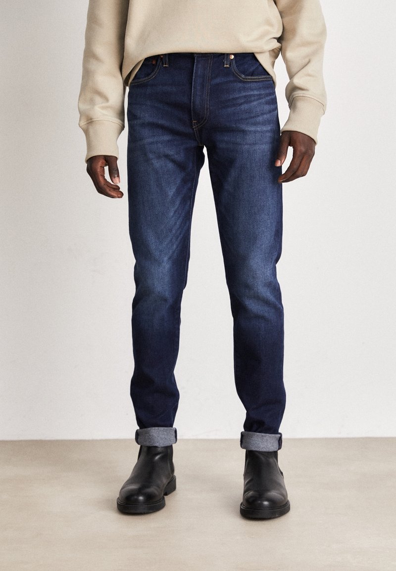 Vaqueros azul oscuro de corte ajustado, con textura de mezclilla, detalles de costuras y puños doblados, combinados con botines negros hasta el tobillo.