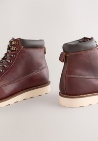 Hnedé kožené členkové čižmy s hladkým povrchom. Majú sivý polstrovaný golier a krémovú gumovú podrážku. Strieborné očká s šnúrkami.