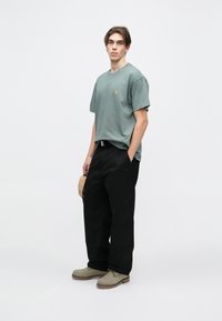 Lys grøn crew-neck t-shirt med logo, sorte brede bukser og grå sko. Model holder en beige taske og står foran en ensfarvet baggrund.