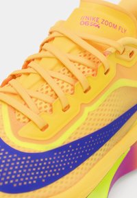 Nike Zoom Fly apavi ir ar spilgtu dzeltenu virsu, kas veidota no sieta materiāla, kam ir oranžas akcenti, zils Swoosh un teksturēta zole rozā un zaļā krāsā.