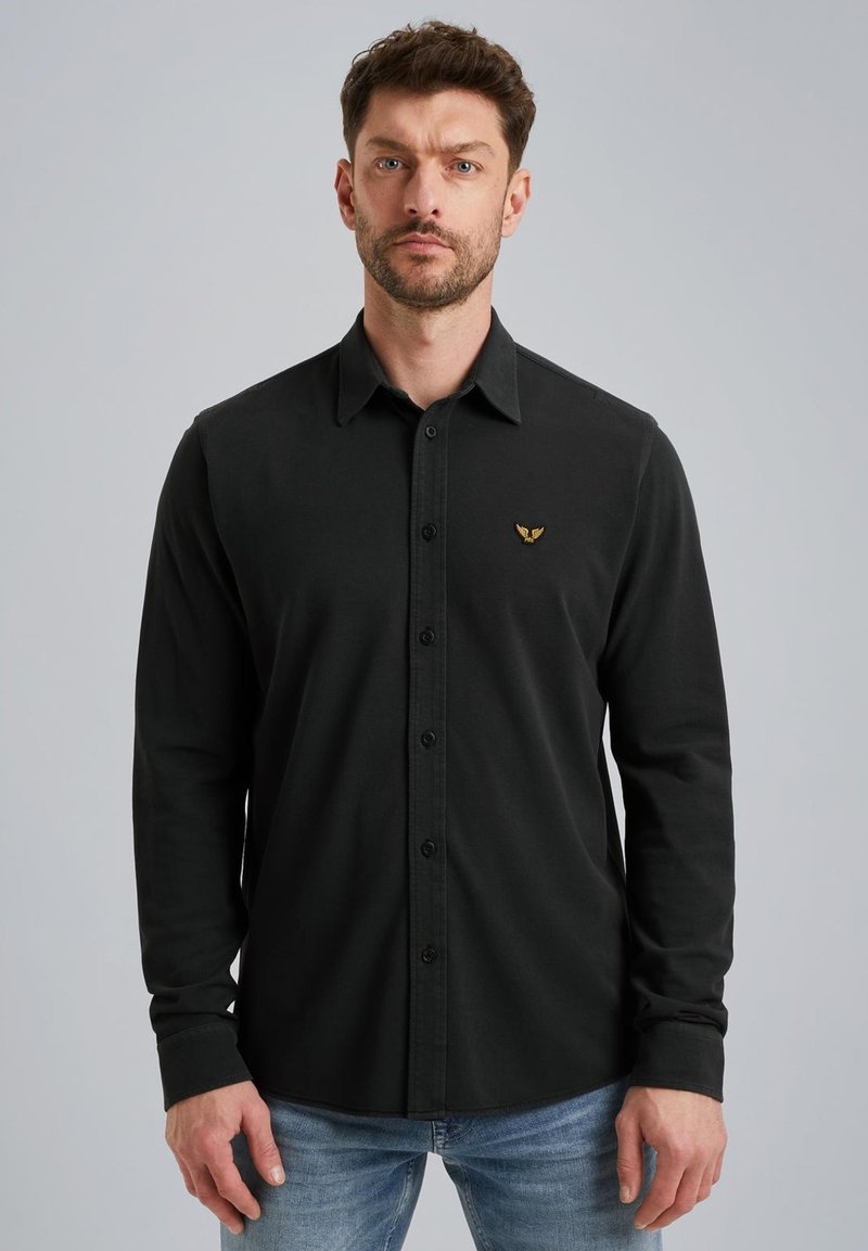 Camisa negra de manga larga con botones y cuello clásico, que presenta un sutil logo dorado en el pecho y una textura de algodón suave.