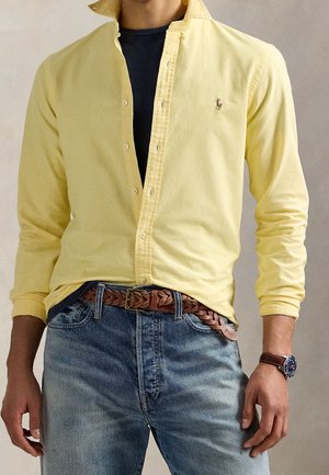 Homme portant une chemise jaune à boutons par-dessus un t-shirt noir, un jean bleu avec une ceinture tressée marron et une montre-bracelet avec un bracelet en cuir.
