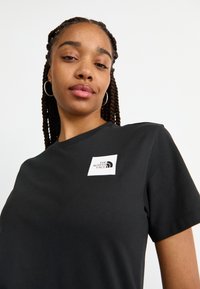 Tricou din bumbac negru cu guler rotund, având un logo pătrat alb pe partea stângă a pieptului cu brandul "The North Face". Textură netedă.