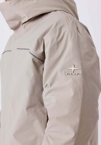 Chaqueta impermeable beige con cuello alto, que presenta una tela sutilmente texturizada, detalles reflectantes y un logo metálico en el hombro.