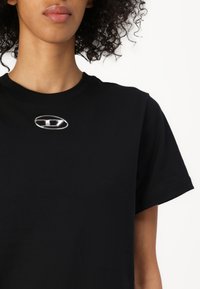 Black t-shirt with a round metallic badge; short sleeves and a crew neckline. Smooth texture, no patterns or additional embellishments.