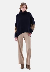 Navy gestrickter Pullover mit hohem Kragen, gemusterte beigen-braunen Ärmel; kombiniert mit beigen, taillierten Hosen und camel-farbenen Ankle Boots.