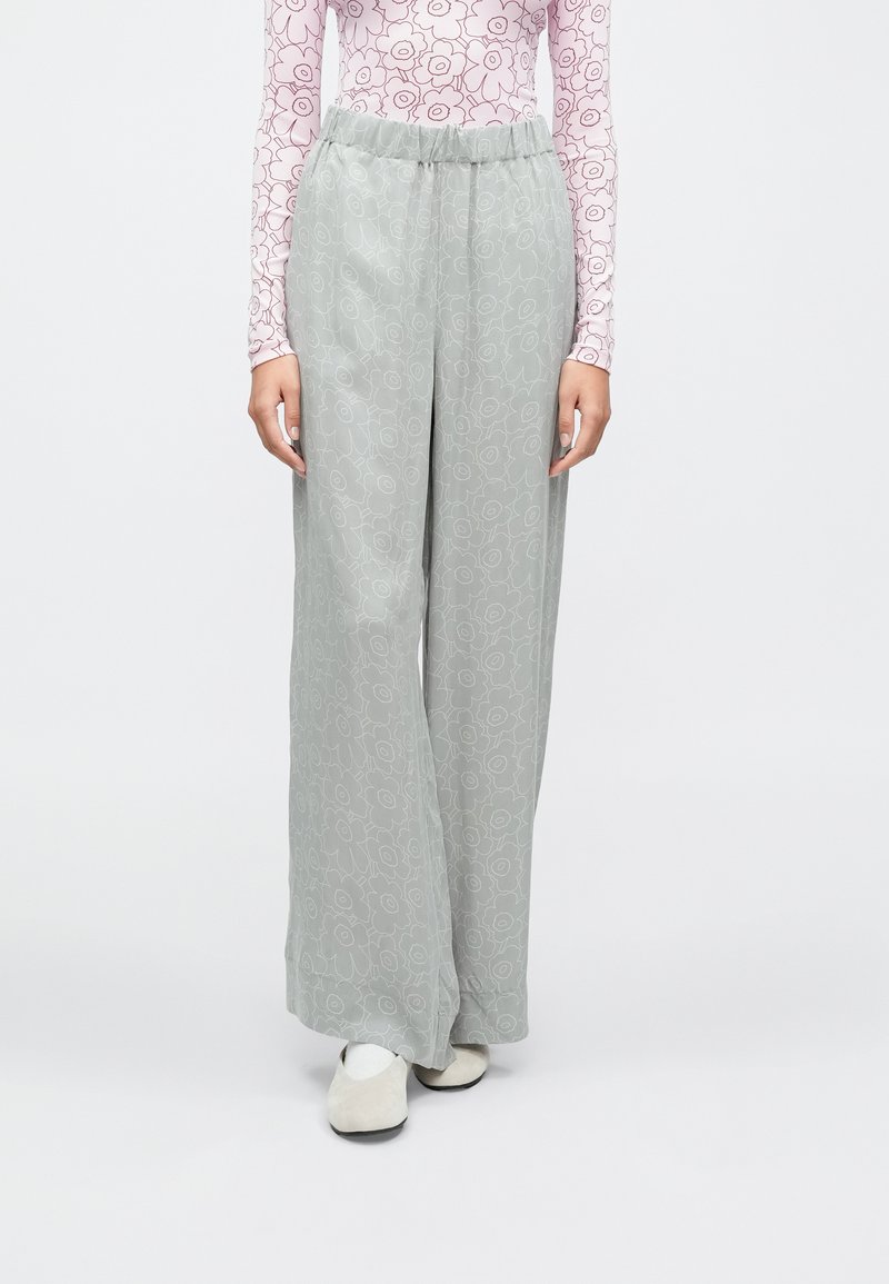 Persona, kas valkā vaļīgas gaiši pelēkas ziedu raksta bikses, rozā ar garām piedurknēm topu ar sarkanu ziedu rakstu, baltas zeķes un bēšas kurpes, stāvot pret baltu fonu.