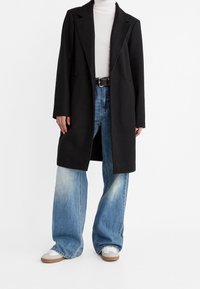 Persoon in een zwarte lange jas, witte coltrui, wijde blauwe jeans, zwarte riem en grijs-witte sneakers tegen een witte achtergrond.