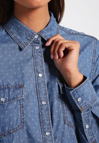 Persona che allaccia il primo bottone di una camicia di jeans blu con un motivo a pois bianchi e tasche frontali.