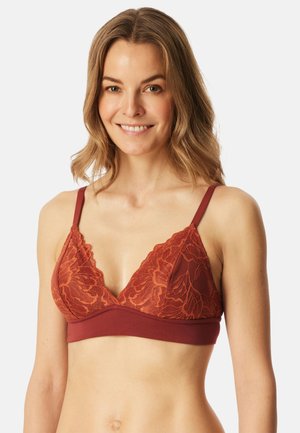 Lächelnde Frau mit leicht welligem Haar, die ein rostfarbenes Spitzen-Bralette mit dünnen verstellbaren Trägern vor einem schlichten Hintergrund trägt.