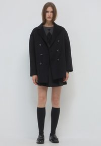 Cappotto nero doppiopetto con bottoni grandi, indossato sopra un maglione grigio a fantasia e una gonna corta; abbinato a calze nere al ginocchio e scarpe.