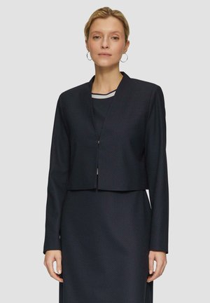 Giacca navy cropped con maniche lunghe, tessuto strutturato, motivo a pois discreto e chiusura frontale. Indossata sopra un vestito abbinato.