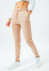 Béžové tepláky s elastickým pasem a stahovací šňůrkou, bočními kapsami, žebrovanými manžetami a bílým logem. Spárované s bílými teniskami.