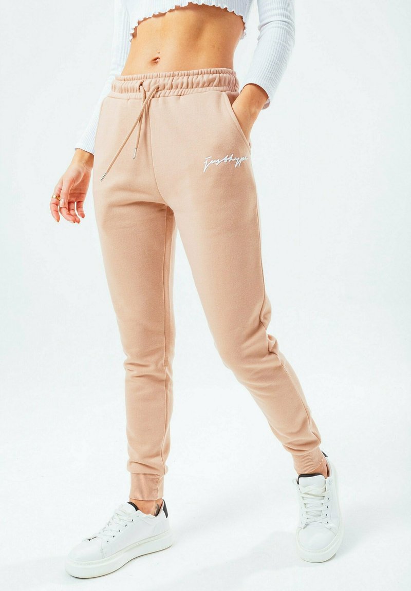 Béžové tepláky s elastickým pasem a stahovací šňůrkou, bočními kapsami, žebrovanými manžetami a bílým logem. Spárované s bílými teniskami.