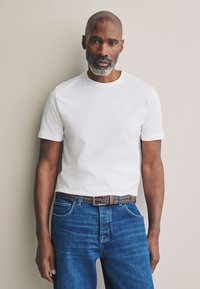 T-shirt bianca a maniche corte abbinata a jeans di denim blu. L'outfit presenta una cintura in pelle intrecciata marrone e elementi di design minimalisti.