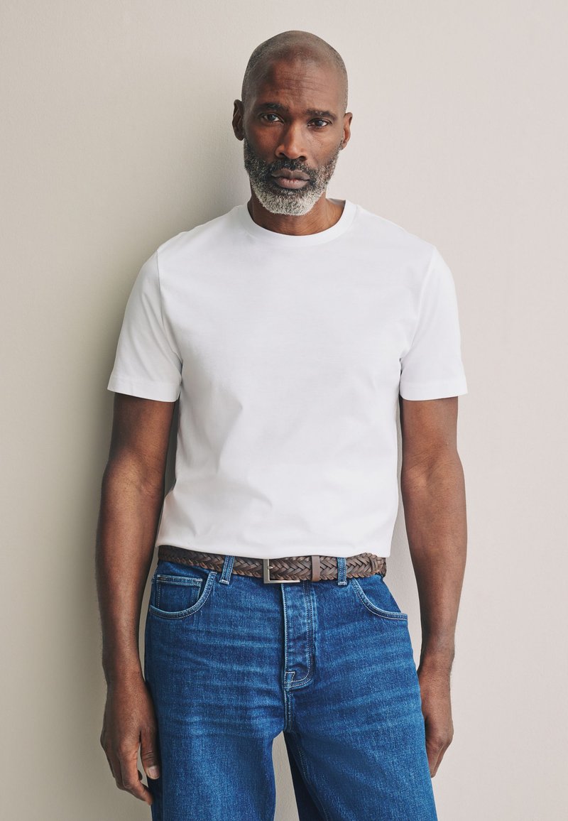 T-shirt bianca a maniche corte abbinata a jeans di denim blu. L'outfit presenta una cintura in pelle intrecciata marrone e elementi di design minimalisti.