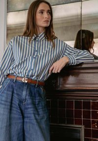 Donna appoggiata su un camino in legno scuro, indossa una camicia a righe verticali blu e bianche infilata in jeans blu larghi con una cintura marrone.